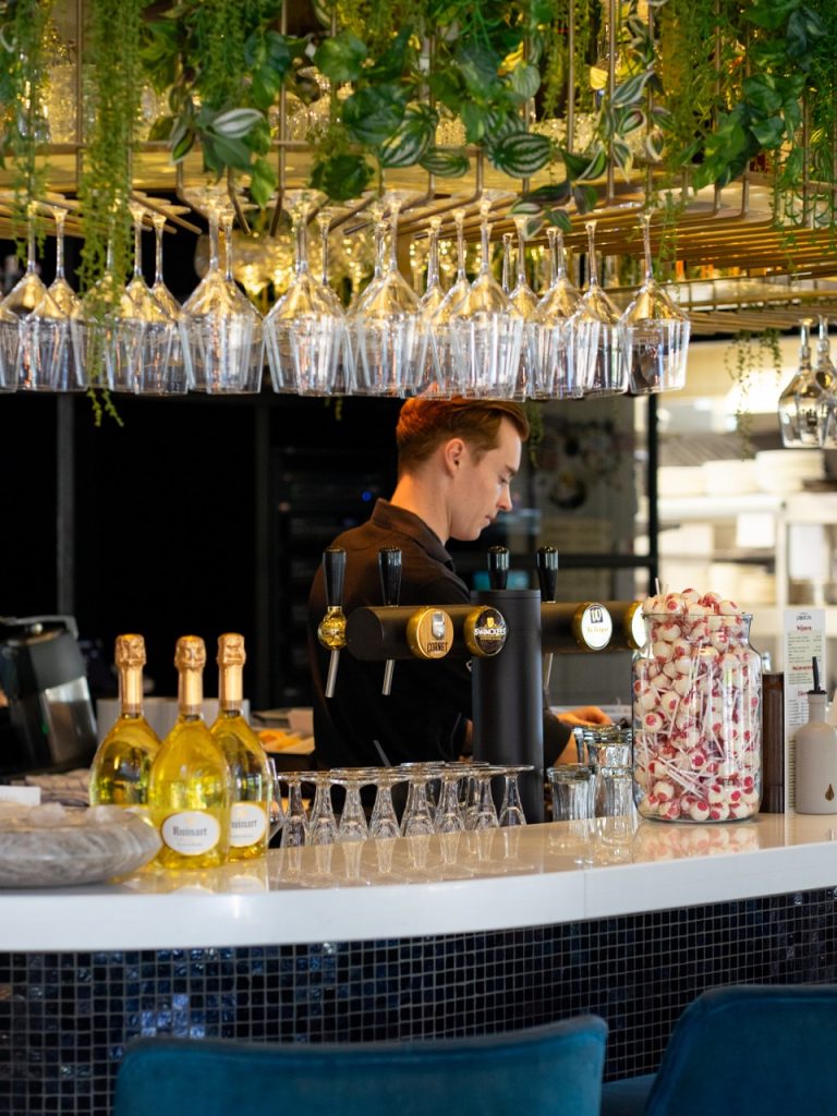 Barman achter de bar van Brasserie Pastis, met champagneflessen en hangende glazen