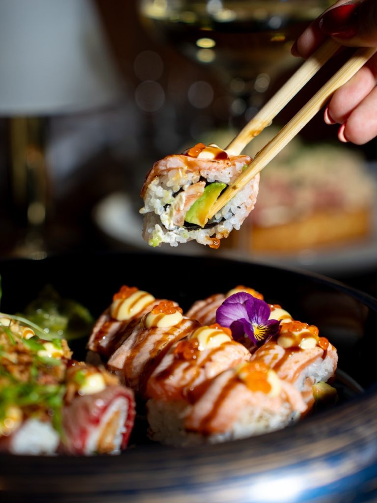 Stokjes pakken een sushiroll met zalm en avocado, op de voorgrond een schaal sushi met een eetbare bloem