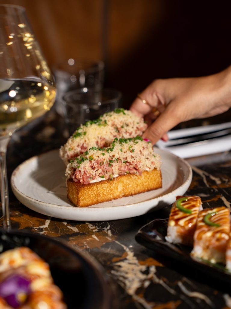 Hand pakt een bordje met belegde brioche, naast sushi op een marmeren tafel