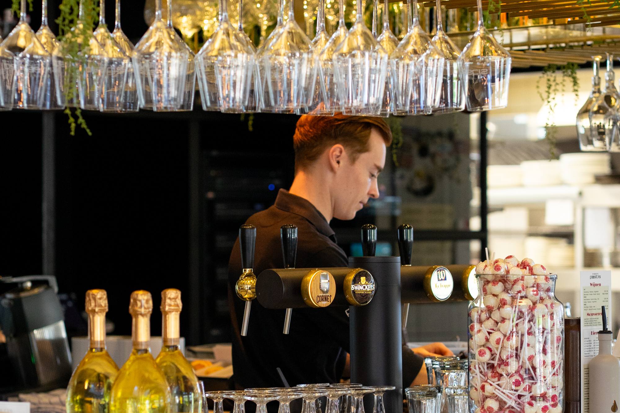 Barman van Brasserie Pastis aan het werk achter de bar, onder het rek met hangende wijnglazen